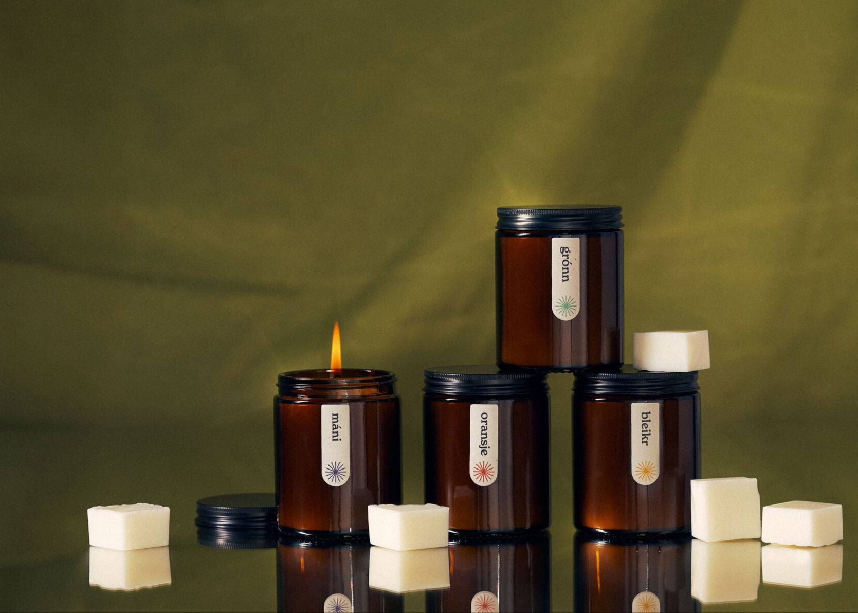 Four amber jar candles labeled máni, grónn, oransje, and bleikr arranged on a reflective surface with white wax cubes