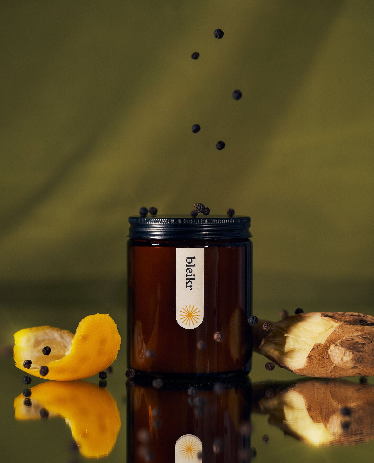 A minimalist amber glass candle labeled “Bleikr,” surrounded by black peppercorns, ginger, and a curled lemon peel, set against a moody green background.