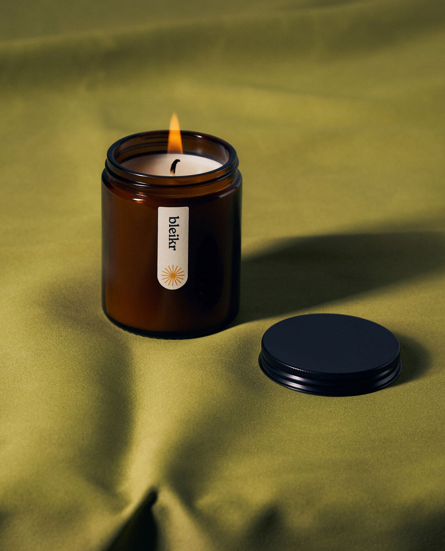 A lit amber glass candle labeled “Bleikr” sits on a textured olive-green fabric, with its black lid placed beside it, casting soft shadows.