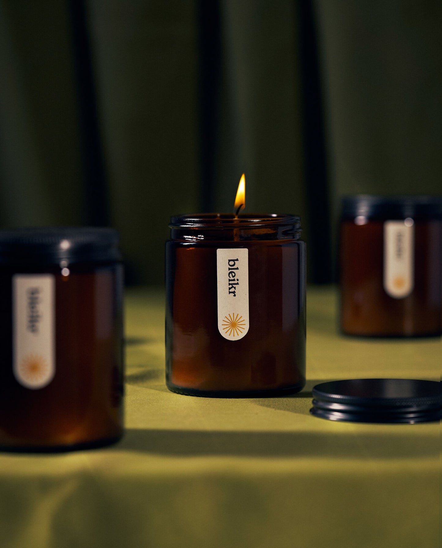 A lit amber jar candle labeled "bleikr" with a minimalist design, surrounded by two unlit candles on a green fabric surface.
