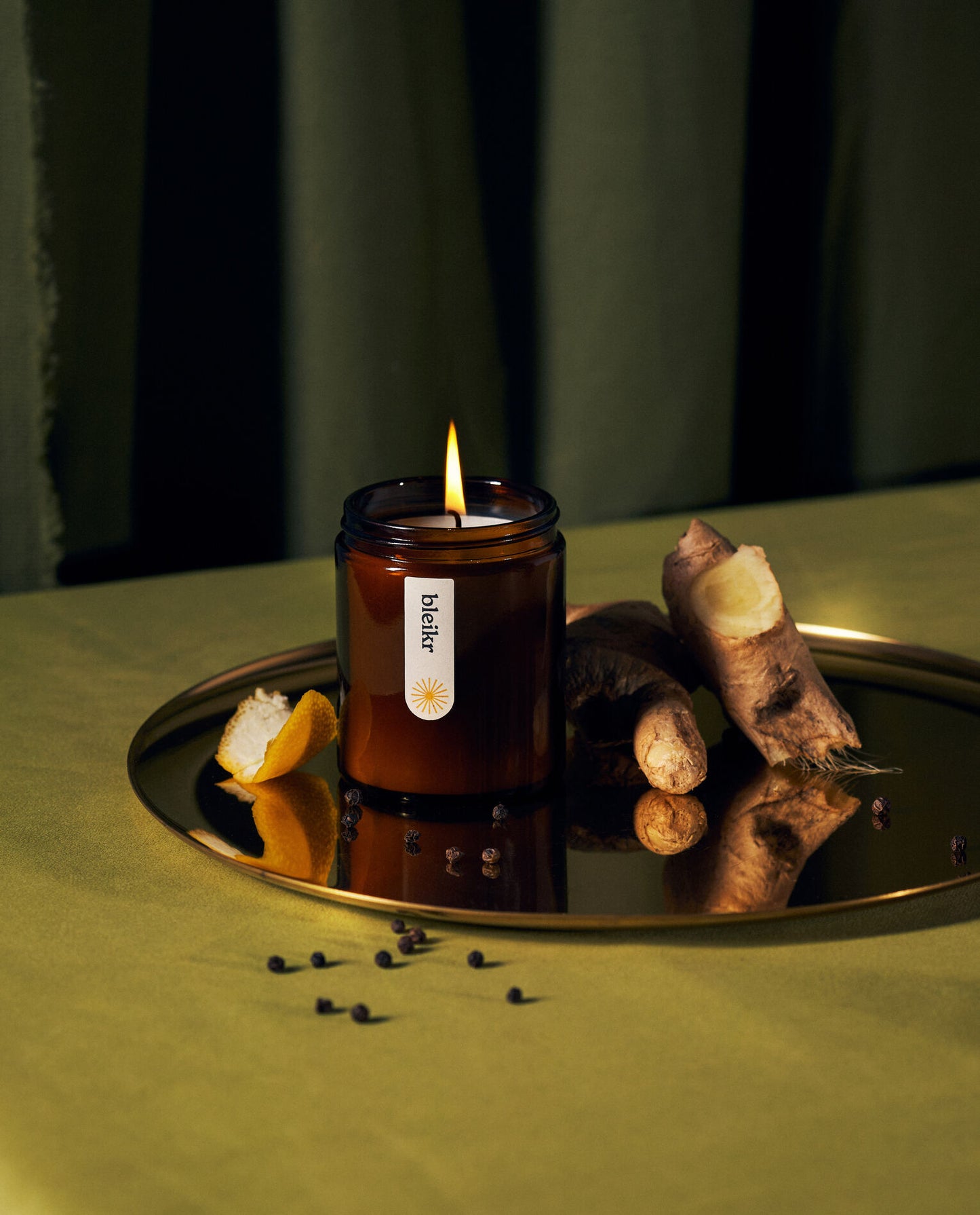 A lit "bleikr" candle on a reflective tray, surrounded by ginger, lemon peel, and scattered peppercorns.