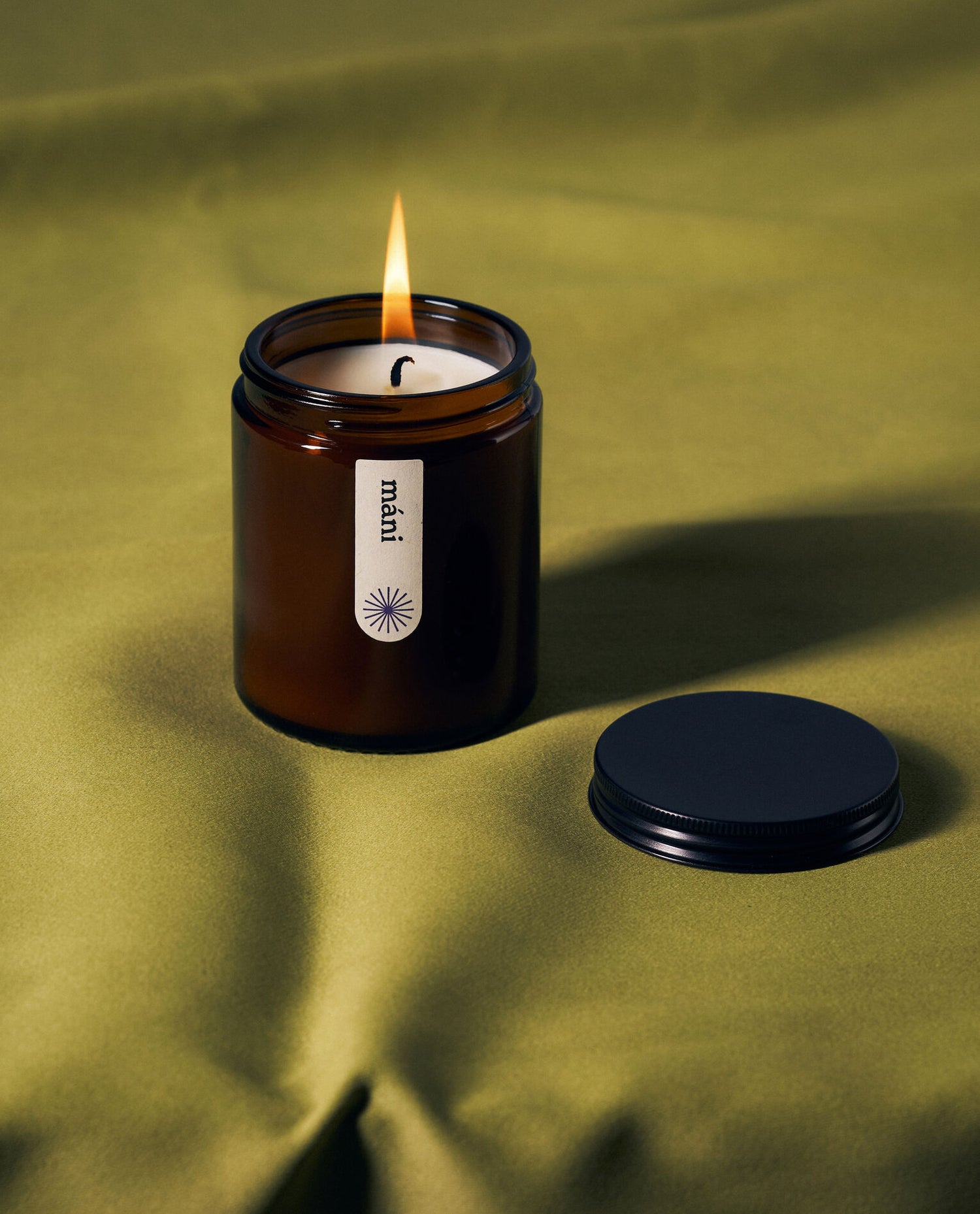 A lit amber jar candle labeled "máni" on green fabric, with its black lid placed nearby.