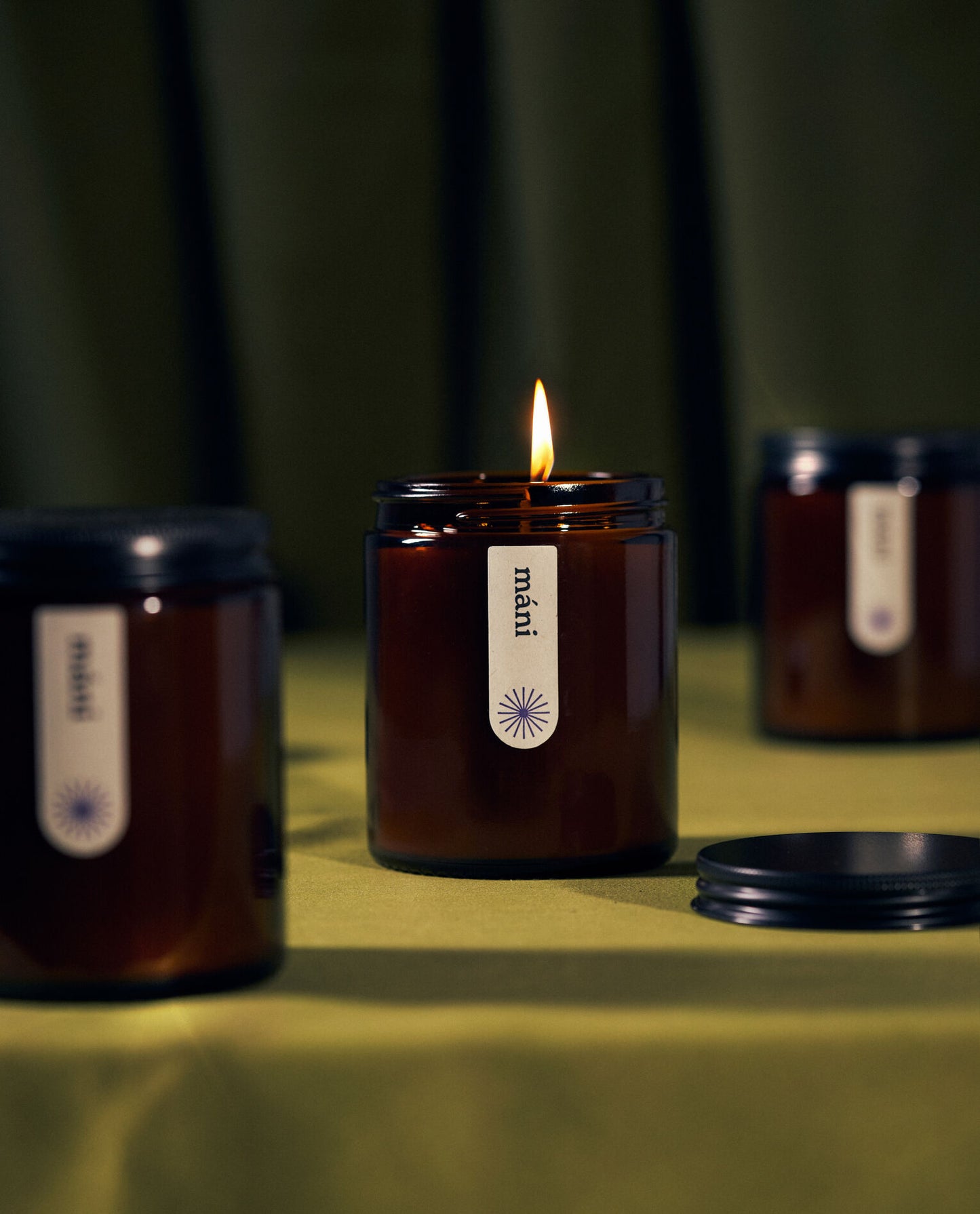 Three amber jar candles labeled "máni" on green fabric, one lit in the centre with black lids scattered around.