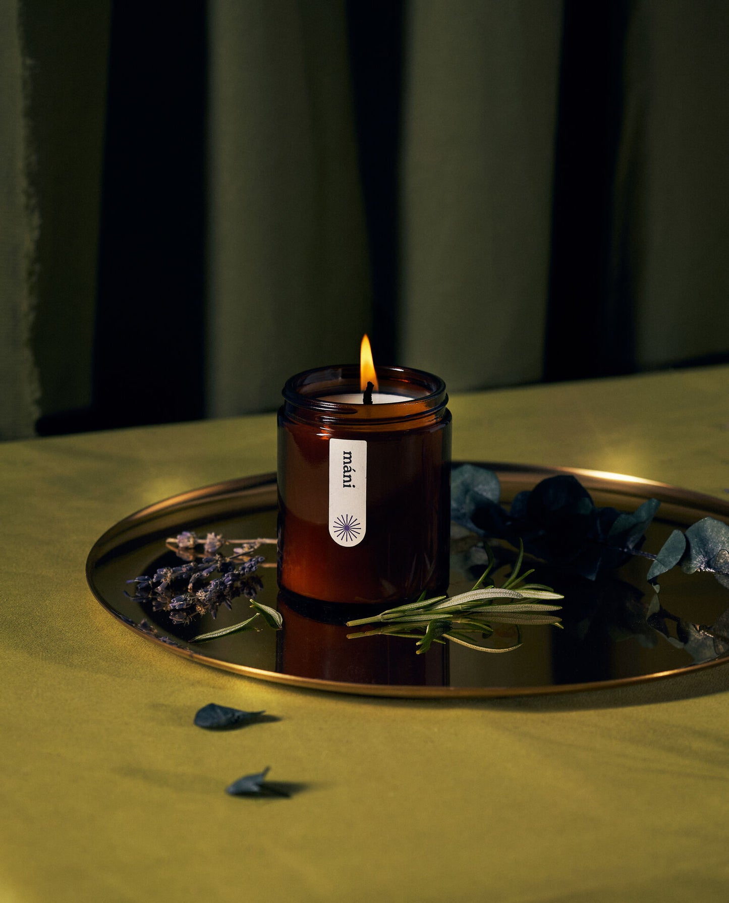 A lit amber jar candle labeled "máni" on a reflective tray, surrounded by lavender sprigs, rosemary, and eucalyptus leaves.