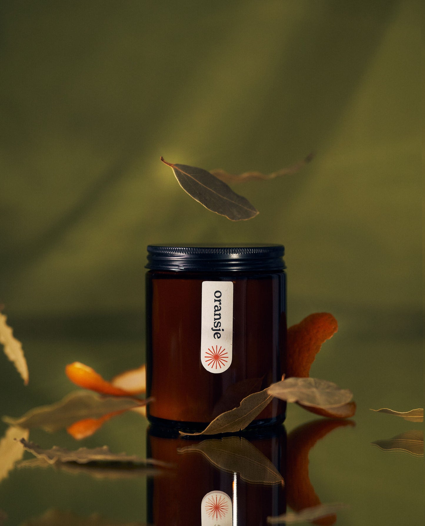 A closed amber jar candle labeled "oransje" with floating bay leaves and orange peel, set on a reflective surface against a green background.