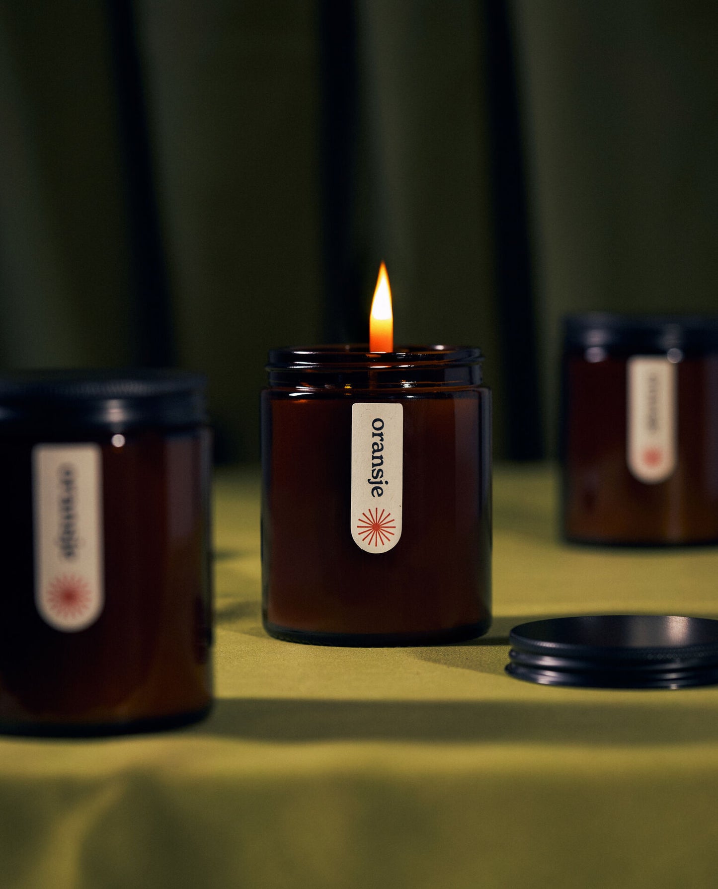 A lit amber jar candle labeled "oransje" on green fabric, surrounded by two unlit candles and a black lid nearby.