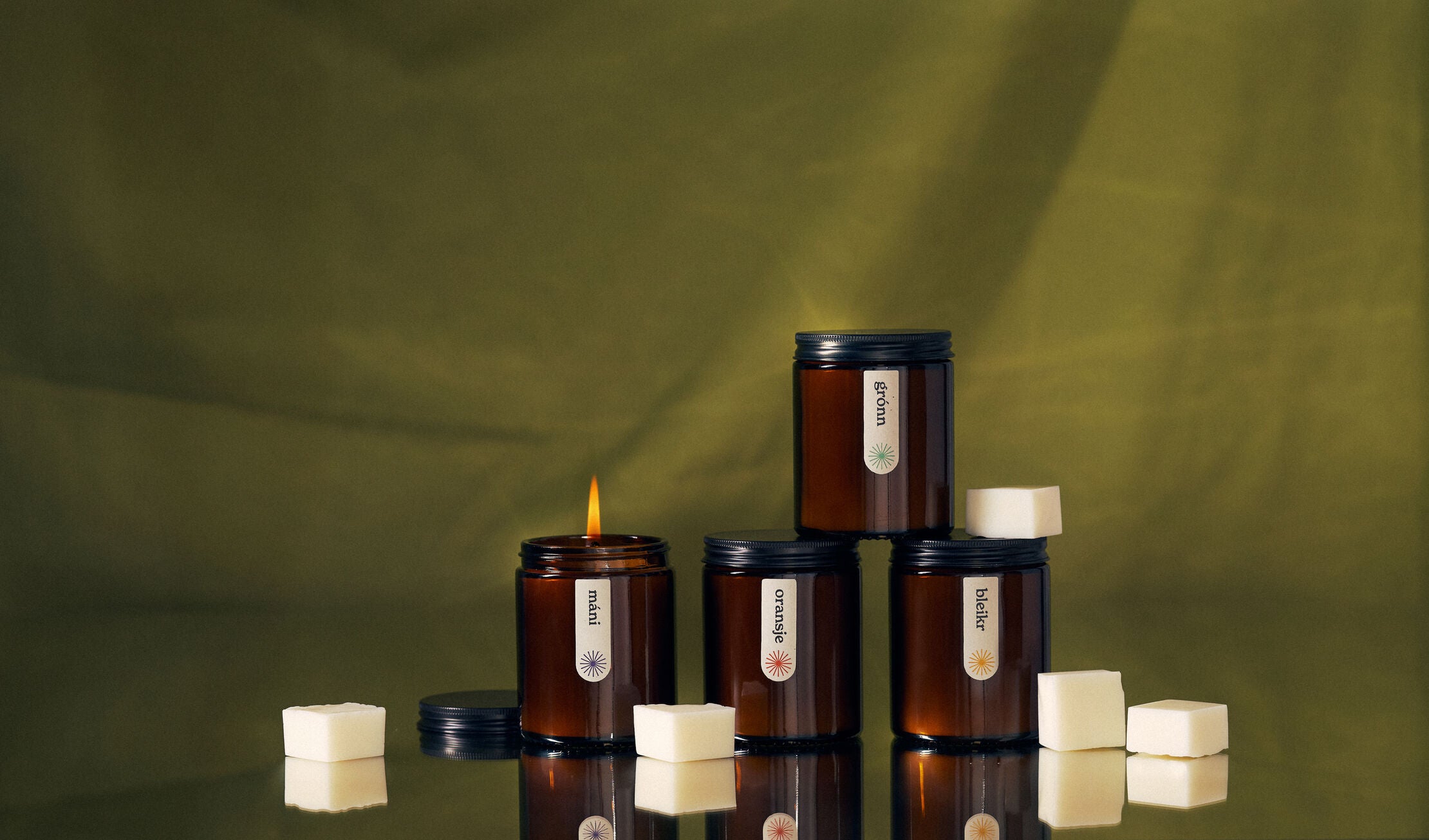 Four amber jar candles labeled "máni," "grónn," "oransje," and "bleikr" arranged on a reflective surface with white wax cubes