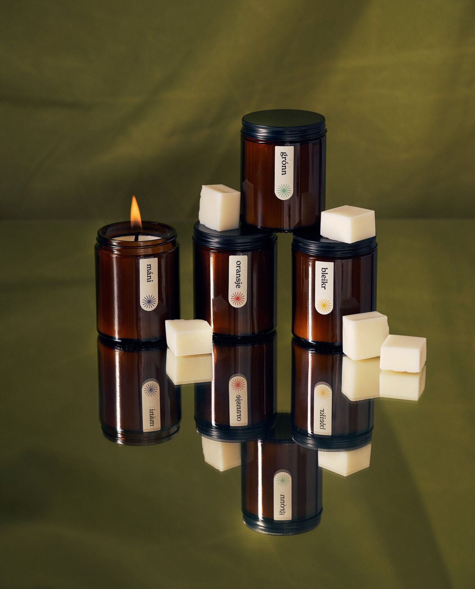 Four amber jar candles labeled "máni," "grónn," "oransje," and "bleikr" arranged on a reflective surface with white wax cubes scattered around, set against a green fabric backdrop.