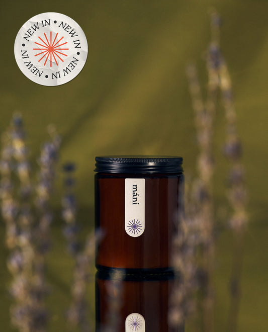 A closed amber jar candle labeled "máni" with a black lid, set against a blurred green background and framed by lavender stems.