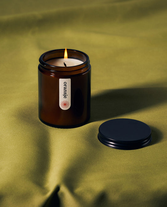 A lit amber jar candle labeled "oransje" on green fabric, with its black lid placed beside it.